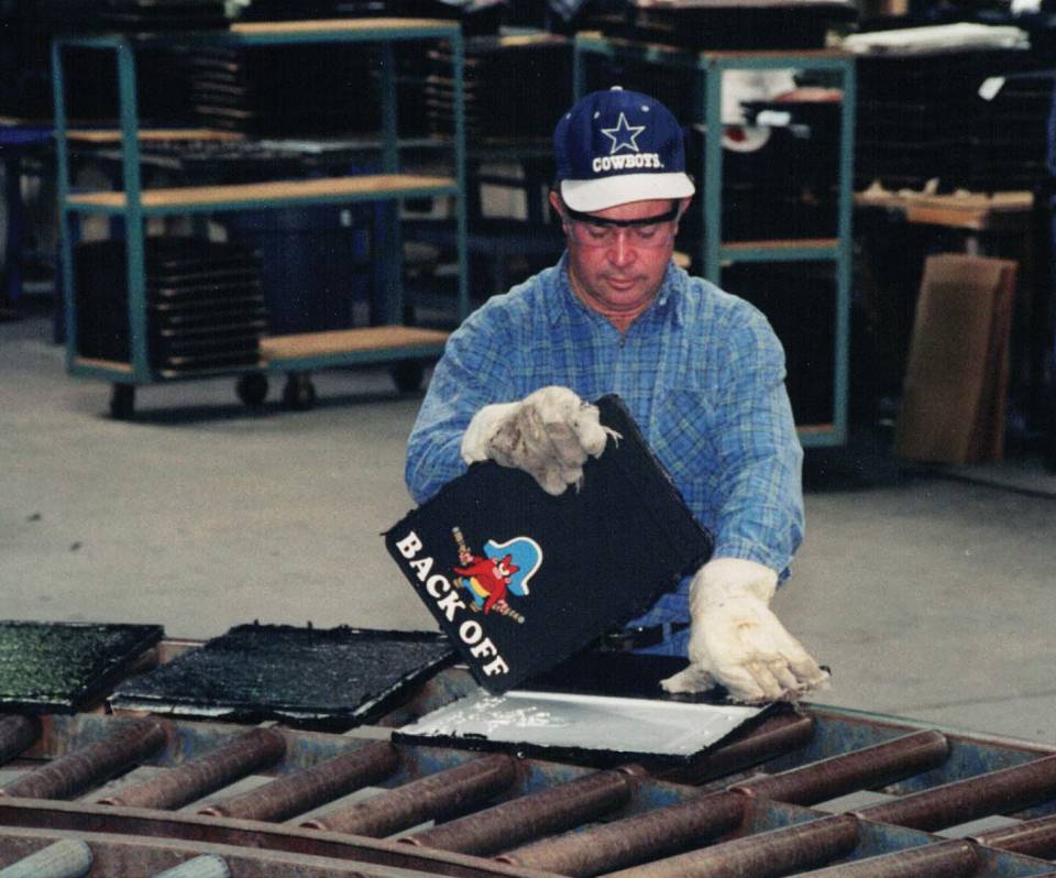 A person in protective gloves inspects a black material with "BACK OFF" printed on it, standing beside industrial rollers in a manufacturing setup.