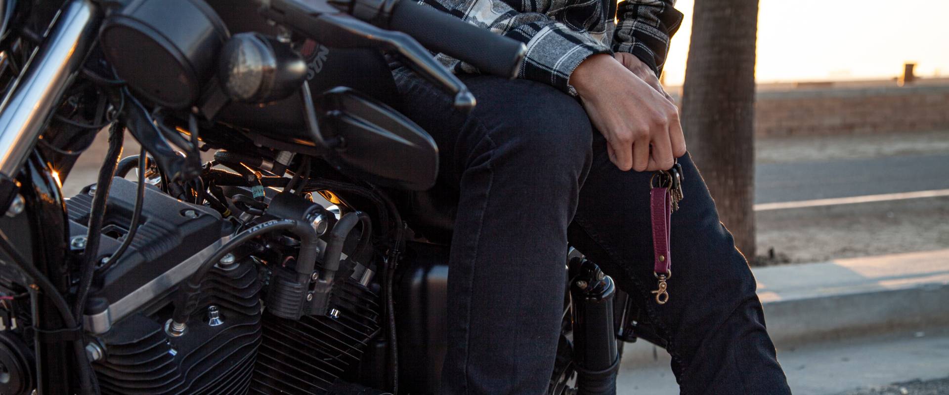 A person is seated on a motorcycle, holding a set of keys, near a road at sunset.