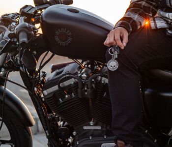 A person is holding a keyring with the Harley-Davidson logo, near a motorcycle with matching branding, during sunset.