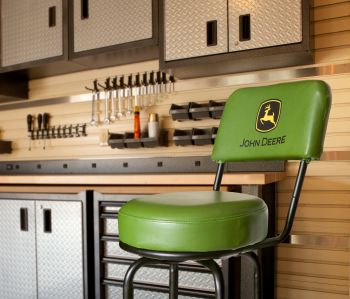 A green John Deere branded bar stool stands in a kitchen with a wooden backsplash, hanging utensils, and cabinetry.