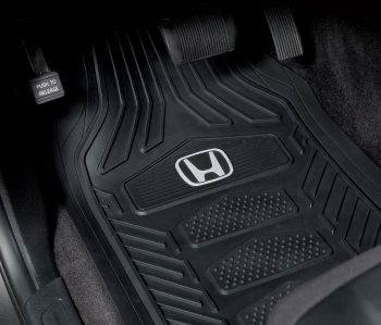 A black car floor mat with a raised Honda emblem rests in a vehicle's footwell, surrounded by pedals and the car's interior trim.