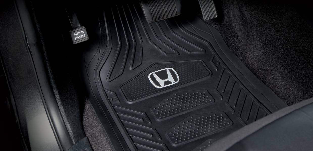 A black, rubber vehicle floor mat with a raised Honda logo, positioned on the interior car floor near pedals, displaying a textured, dirt-resistant design.