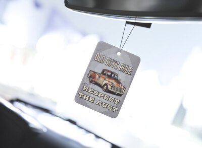 A square air freshener hangs inside a vehicle, bearing the illustration of a vintage truck and the text "OLD UNSPOILED - RESPECT THE RUST." The background is softly blurred.