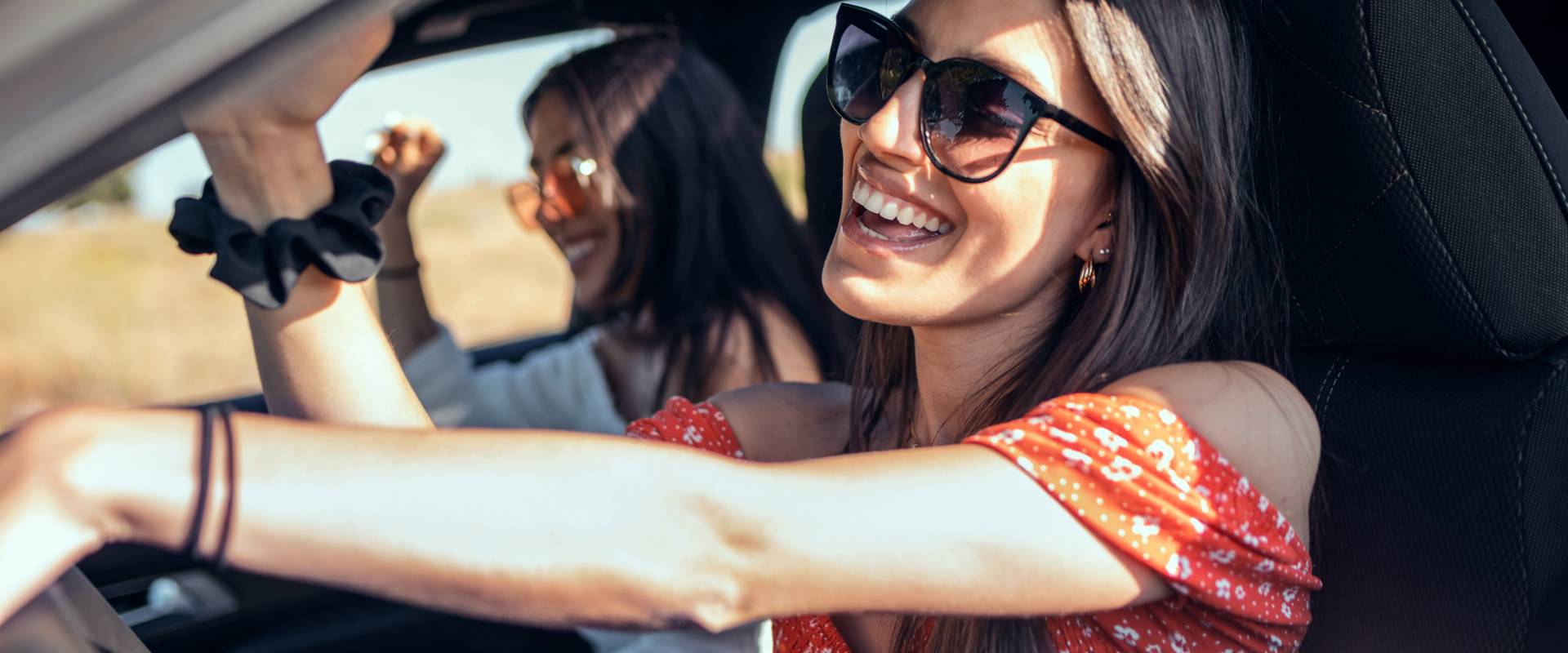 A smiling woman wearing sunglasses is driving a car, with a passenger raising her arms joyfully in the background, all against a sunny outdoor backdrop.