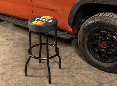 A round stool with a black metal frame and a colorful striped cushion is placed near an orange car's black wheel on a concrete floor.