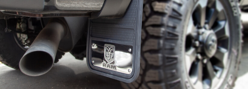 A black exhaust tip protrudes from a vehicle's side step, adorned with a "RAM" logo badge, near a rugged tire, indicating a heavy-duty truck.