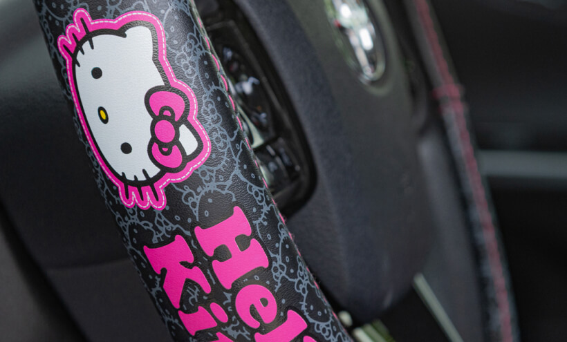 A Hello Kitty character decorates a steering wheel cover, accented with a pink logo and designs, inside a vehicle.