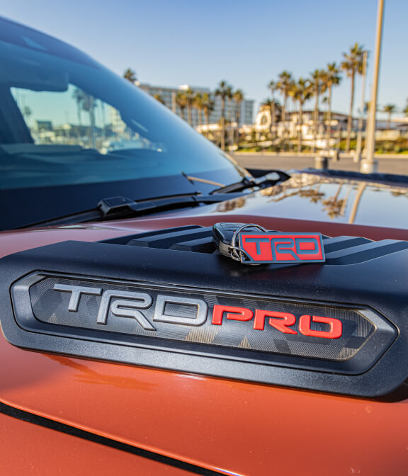 A red "TRD PRO" badge on a vehicle's hood with palm trees and a blue sky in the background, suggesting a sunny outdoor setting.