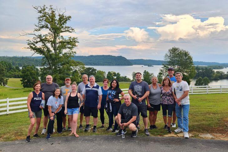 A group of adults posing and smiling for a group photo with a scenic lake and greenery in the background.