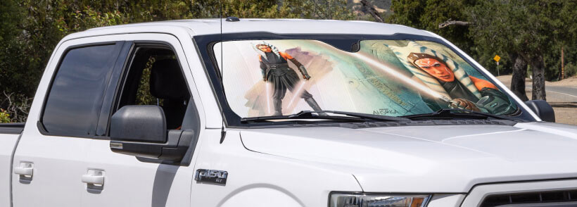 A white pickup truck features a windshield sunshade with an illustration of a female character in an action pose, parked in a sunny, outdoor setting.